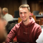 Ein Mann mit dunklen Haaren und einem Anzug sitzt an einem Pokertisch und betrachtet konzentriert seine Karten.