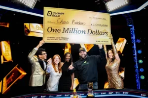 Ein Mann sitzt an einem Pokertisch und hält Karten in der Hand. Er trägt eine schwarze Kappe und ein schwarzes Hemd. Im Hintergrund sind weitere Personen und Pokerchips zu sehen.