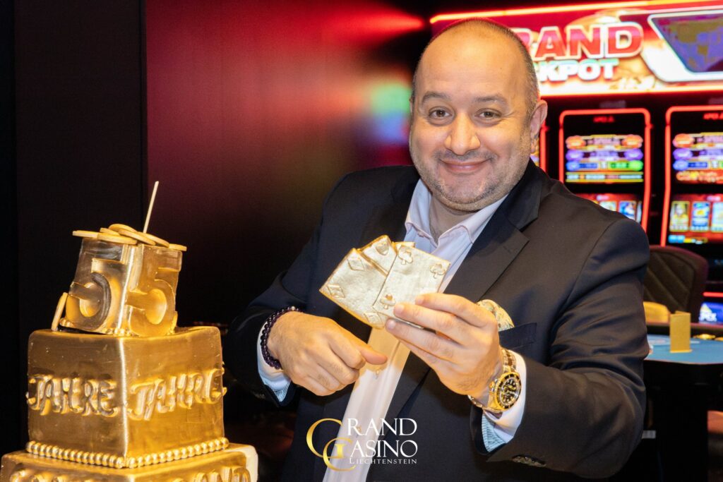 Ein eleganter Pokertisch mit grünem Filz und einem Stapel von Jetons, umgeben von Spielkarten und Gläsern. Menschen in festlicher Kleidung sitzen um den Tisch herum und genießen