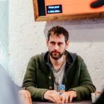 Ein älterer Mann in einem dunkelblauen Anzug sitzt an einem Pokertisch und hält Pokerkarten in der Hand. Er hat graue Haare, trägt eine Brille und schaut konzentriert auf