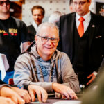 Ein älterer Mann, Herbert Gottsbacher, sitzt lächelnd an einem Pokertisch und hält Karten in der Hand. Er trägt ein dunkles Hemd und eine Brille. Im Hintergrund sind weitere