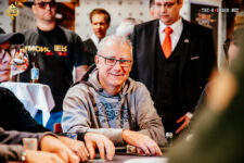 Ein älterer Mann, Herbert Gottsbacher, sitzt lächelnd an einem Pokertisch und hält Karten in der Hand. Er trägt ein dunkles Hemd und eine Brille. Im Hintergrund sind weitere