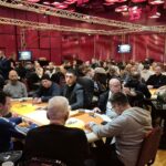 Ein Mann spielt Poker an einem Tisch mit grüner Filzdecke. Er hält Karten in der Hand und schaut konzentriert auf den Tisch. Im Hintergrund sind Chips und Getränke zu