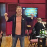 Ein Mann in einem schwarzen Anzug und einer roten Krawatte sitzt an einem Pokertisch und hält zwei Könige (King of Hearts und King of Diamonds) in der Hand. Er hat einen ernsten Gesichtsa