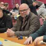 Ein professioneller Pokertisch mit grüner Filzoberfläche, umgeben von Stühlen und Chips. In der Mitte des Tisches befinden sich Spielkarten und Spieler, die ihre Hände überprüfen.