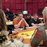 Ein professioneller Pokertisch mit grüner Filzoberfläche, umgeben von gepolsterten Stühlen. Auf dem Tisch liegen Spielkarten, Pokerchips und Getränke. Im Hintergrund