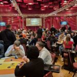 Ein Mann in einem dunklen Anzug und einer schwarzen Sonnenbrille sitzt an einem Pokertisch mit grünem Filz. Er hält seine Karten in der Hand und schaut konzentriert auf den Tisch