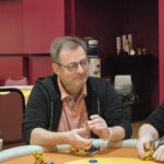 Ein Bild eines Pokertisches mit Spielkarten, Pokerchips und Getränken, umgeben von Spielern in einem Casino.