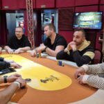 Ein professioneller Pokertisch mit grüner Filzoberfläche, umgeben von mehreren Spielern, die Karten halten und Chips stapeln. Ein Dealer sitzt in der Mitte und mischt die Karten. Die Spieler