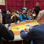 Ein Mann sitzt an einem Pokertisch und hält zwei Karten in der Hand. Er trägt eine dunkle Brille und konzentriert sich auf das Spiel. Um ihn herum liegen Pokerchips und Karten auf