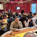 Ein Pokertisch mit grüner Filzdecke, umgeben von Spielkarten, Pokerchips und Getränken. Vier Personen sitzen um den Tisch herum, konzentriert auf ihre Karten.