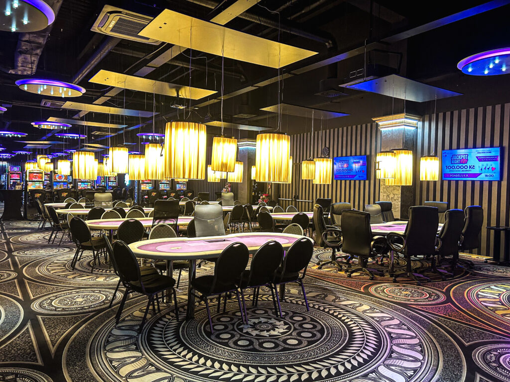 Ein Mann sitzt an einem Pokertisch in einem Casino. Er hat eine Hand mit Pokerchips und eine Karte in der Hand. Im Hintergrund sind andere Spieler und Dealer zu sehen.