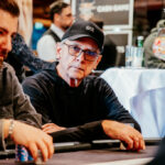 Ein Mann mit dunklen Haaren und einem Bart, der konzentriert an einem Pokertisch sitzt und Karten in der Hand hält. Er trägt ein dunkles Hemd und eine schwarze Kappe. Im Hinter