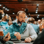 Ein Mann und eine Frau sitzen an einem Pokertisch und halten ihre Karten. Der Mann hat eine ernste Miene, während die Frau lächelt. Im Hintergrund sind Pokerchips und Getränke zu sehen.