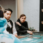 Ein Mann in einem schwarzen Anzug und einer roten Krawatte sitzt an einem Pokertisch und hält Karten in der Hand, während er konzentriert auf seine Gegner schaut.