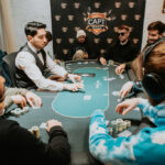 Ein Mann in einem dunkelblauen Anzug sitzt an einem Pokertisch und hält zwei Spielkarten in der Hand. Er schaut konzentriert auf seine Karten, während andere Spieler im Hintergrund zu sehen sind