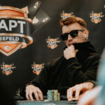 Ein Mann in einem dunkelblauen Anzug sitzt an einem Pokertisch und hält eine Hand mit Spielkarten. Er schaut konzentriert auf seine Karten, während im Hintergrund andere Spieler und Chips zu sehen