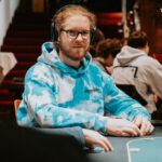Ein Mann sitzt an einem Pokertisch und hält eine Hand mit Spielkarten. Er trägt einen dunklen Anzug und eine Krawatte. Um ihn herum sind Pokerchips und Getränke zu sehen.