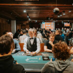 Ein Mann sitzt an einem Pokertisch, hält Karten in der Hand und betrachtet sie konzentriert, umgeben von anderen Spielern und Pokerchips.