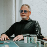 Ein Mann sitzt an einem Pokertisch und hält zwei Karten in der Hand, während er konzentriert auf die Gemeinschaftskarten in der Mitte des Tisches schaut. Andere Spieler sind im Hintergrund