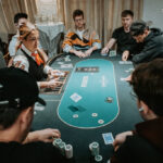 Ein Mann sitzt an einem Pokertisch und hält zwei Spielkarten in der Hand, während er konzentriert auf die Mitte des Tisches schaut. Andere Spieler sind im Hintergrund zu sehen.