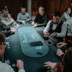 Ein Mann sitzt an einem Pokertisch und hält zwei Spielkarten in der Hand. Er wirkt konzentriert und trägt eine Kappe sowie einen dunklen Pullover. Neben ihm liegen Pokerchips und eine