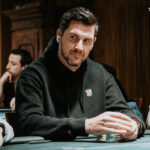 Ein Mann in einem dunkelblauen Anzug sitzt an einem Pokertisch und hält in einer Hand Spielkarten. Er schaut konzentriert auf seine Karten und hat eine ernste Miene. Im Hintergrund