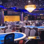 Ein Mann sitzt an einem Pokertisch und hält zwei Spielkarten in der Hand. Er schaut konzentriert auf den Tisch, wo Pokerchips und weitere Karten liegen. Im Hintergrund sind weitere Spieler