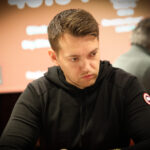 Ein Mann sitzt an einem Pokertisch mit grünen Filz und hält zwei Asse in der Hand. Er hat eine ernste Miene und trägt einen dunklen Anzug. Im Hintergrund sind weitere Spieler und Chips