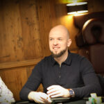 Ein Mann und eine Frau sitzen an einem Pokertisch und spielen Karten. Der Mann hat einen Stapel Chips vor sich und sieht konzentriert aus, während die Frau ihre Karten betrachtet. Im Hintergrund sind weitere