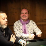 Ein professioneller Pokerspieler mit Sonnenbrille und Kappe sitzt an einem Pokertisch mit grüner Filzdecke. Er betrachtet konzentriert seine Karten während er in einer entspannten H