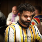 Ein Mann und eine Frau sitzen an einem Pokertisch und schauen konzentriert auf ihre Karten. Der Mann trägt einen dunklen Anzug, die Frau ein elegantes Kleid. Im Hintergrund sind Dealer und andere