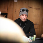 Ein glücklicher Mann in einem schwarzen Anzug und einer Fliege, der lächelnd eine Handkarte mit Pik-As und Pik-König hält, während er an einem Pokertisch sitzt. Im H