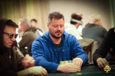 Ein professioneller Pokerspieler mit einer Sonnenbrille und einem schwarzen Hut, der konzentriert an einem Pokertisch sitzt und Karten hält, umgeben von anderen Spielern und Chips.