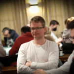 Ein Mann mit dunkler Hautfarbe und einem blauen Anzug, der Poker spielt und konzentriert auf seine Karten schaut. Er sitzt an einem grünen Pokertisch mit Chips und anderen Spielern im H
