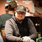 Ein Mann in einem schwarzen Anzug und einer weißen Hemdweste sitzt an einem Pokertisch und hält eine Hand mit Spielkarten. Er wirkt konzentriert und fokussiert. Im Hintergrund