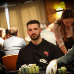 Ein Mann in einem schwarzen Anzug sitzt an einem Pokertisch mit grünem Filz. Er hält Spielkarten in der Hand und schaut konzentriert auf den Tisch. Um ihn herum sind Pokerch