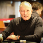 Ein Mann in schwarzer Kleidung und Sonnenbrille sitzt an einem Pokertisch und betrachtet konzentriert seine Karten. Im Hintergrund sind weitere Spieler und Chips zu sehen.