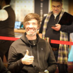 Ein Mann mit dunklen Haaren und einem schwarzen Anzug sitzt an einem Pokertisch und hält zwei Spielkarten in der Hand. Ein Stapel Pokerchips liegt vor ihm auf dem grünen Filz. Im H