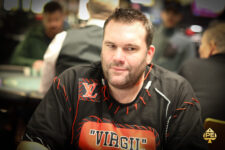Ein Mann sitzt an einem Pokertisch und hält seine Karten mit einem konzentrierten Gesichtsausdruck. Um ihn herum stehen weitere Spieler und beobachten das Spielgeschehen. Die Atmosphäre ist