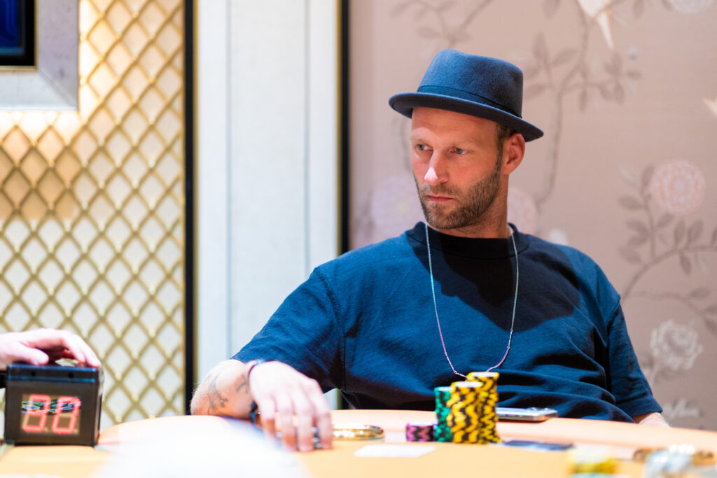 Ein Mann sitzt an einem Pokertisch und hält seine Karten. Im Hintergrund sind weitere Spieler und Chips zu sehen.
