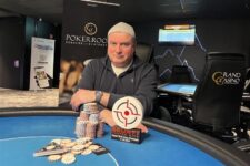 Ein Spieler hält drei Spielkarten in der Hand und schaut konzentriert auf den Tisch, der mit Pokerchips und weiteren Karten gefüllt ist. Es handelt sich um eine Szene aus dem Pok