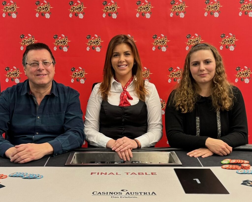 Ein Mann und eine Frau sitzen an einem Pokertisch und spielen Karten. Der Mann hat eine Hand voll Spielchips, während die Frau mit ernstem Blick ihre Karten betrachtet. Im Hintergrund sind weitere Personen und eine