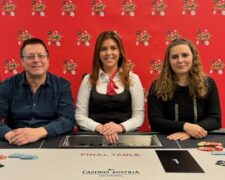 Ein Mann und eine Frau sitzen an einem Pokertisch und spielen Karten. Der Mann hat eine Hand voll Spielchips, während die Frau mit ernstem Blick ihre Karten betrachtet. Im Hintergrund sind weitere Personen und eine