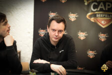Ein Mann sitzt an einem Pokertisch mit Chips und Karten vor sich. Er hat eine ernste Miene und trägt ein dunkles Hemd.