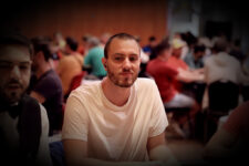 Ein Mann sitzt an einem Pokertisch und betrachtet konzentriert seine Karten. In der Hand hält er Pokerchips und trägt eine schwarze Kappe. Im Hintergrund sind verschwommene Spieler und