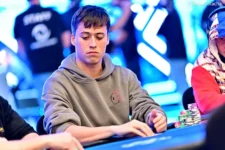 Ein Mann mit dunklen Haaren und einem Bart sitzt an einem Pokertisch und hält seine Karten in der Hand. Er schaut konzentriert auf den Tisch. Im Hintergrund sind andere Spieler und Pokerchips