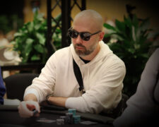 Ein Mann sitzt lächelnd an einem Pokertisch und hält Karten in der Hand. Er trägt ein schwarzes Hemd und hat kurze braune Haare. Im Hintergrund sind Pokerchips und andere Spieler