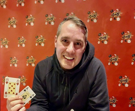 Ein Mann spielt Poker an einem Tisch in einem eleganten Casino. Er hat einen ernsten Gesichtsausdruck und hält Karten in der Hand, während er von anderen Spielern umgeben ist. Die Szene strah