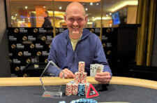 Ein Mann mit dunklem Haar und Bart, der eine Sonnenbrille trägt und eine Pokerkarte in der Hand hält, während er konzentriert in die Kamera schaut.
